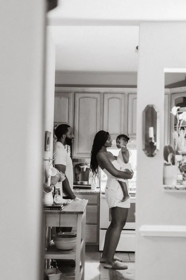 Black and white photo of a family in a kitchen, mother holding baby while father stands nearby.