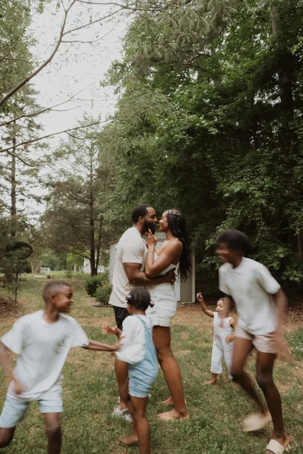 Loving Black couple embracing outdoors while their three children play around them in a natural setting.