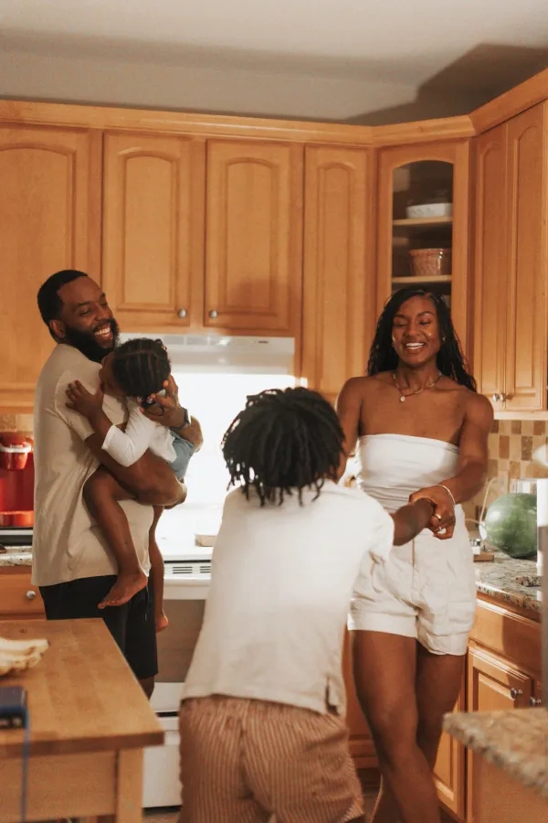 Happy Black family laughing and playing in their kitchen, father holding young child, mother holding hands with older child.