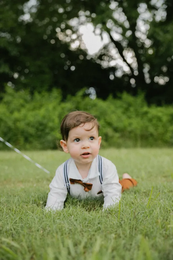 Cute baby boy in white shirt and suspenders crawling on green grass, looking up, outdoor portrait.