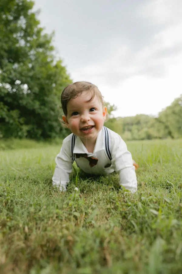 Smiling baby in white shirt and bow tie crawling on green grass outdoors, trees in background.