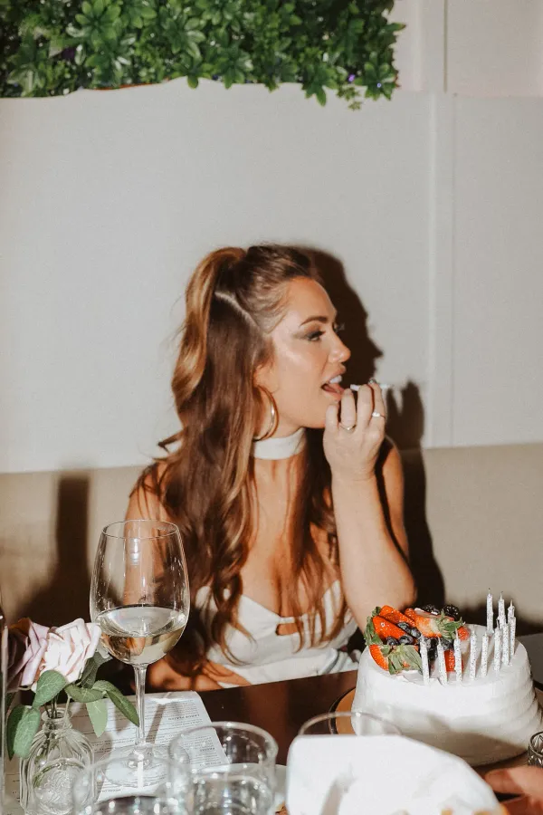 Woman celebrating birthday with cake and wine at a restaurant table, enjoying a fun moment.