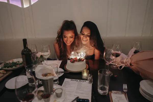 Two smiling women blowing out candles on a birthday cake at a restaurant table in Charlotte, NC.