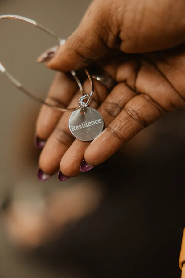 Close-up of a hand holding a silver bracelet with a 'Resilience' charm, purple nails visible.