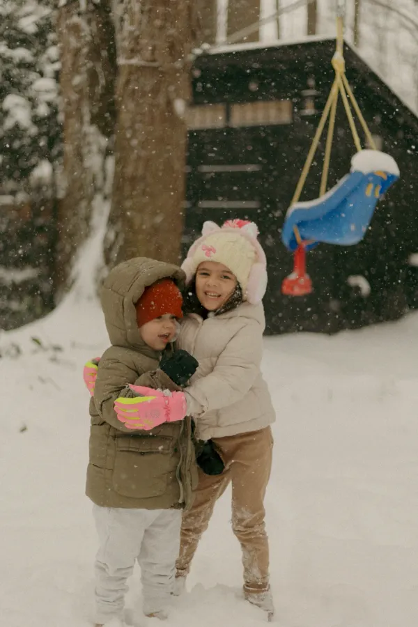 Two smiling children in winter coats and hats hugging in a snowy backyard, snow falling around them.