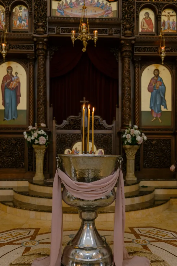 Ornate church interior with a silver baptismal font, pink fabric, and lit candles, surrounded by religious icons.