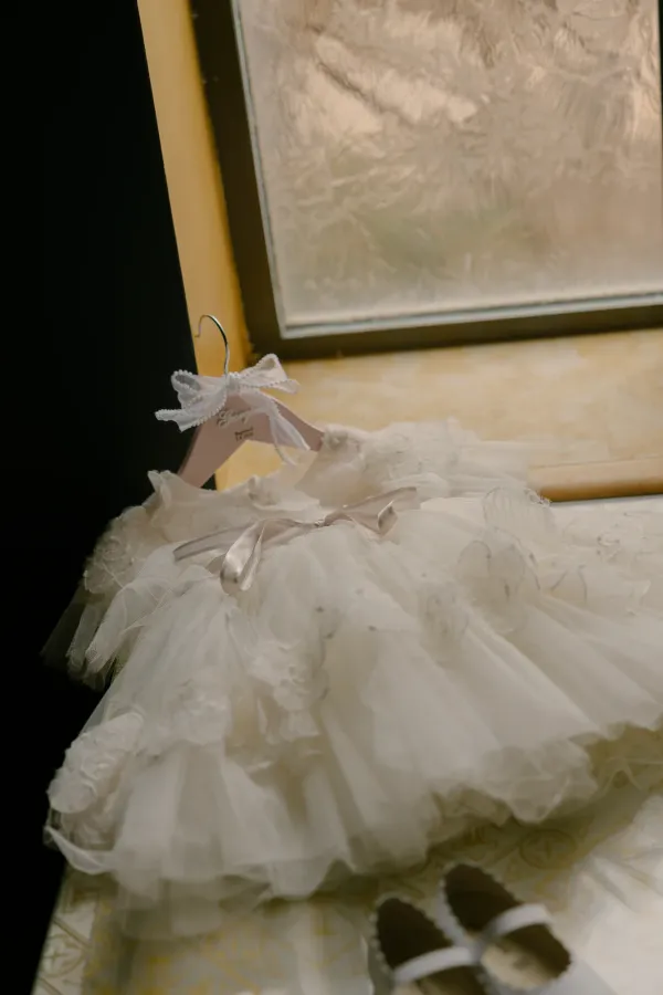 White baby christening dress with lace and ribbons, tiny white Mary Jane shoes, next to a window.