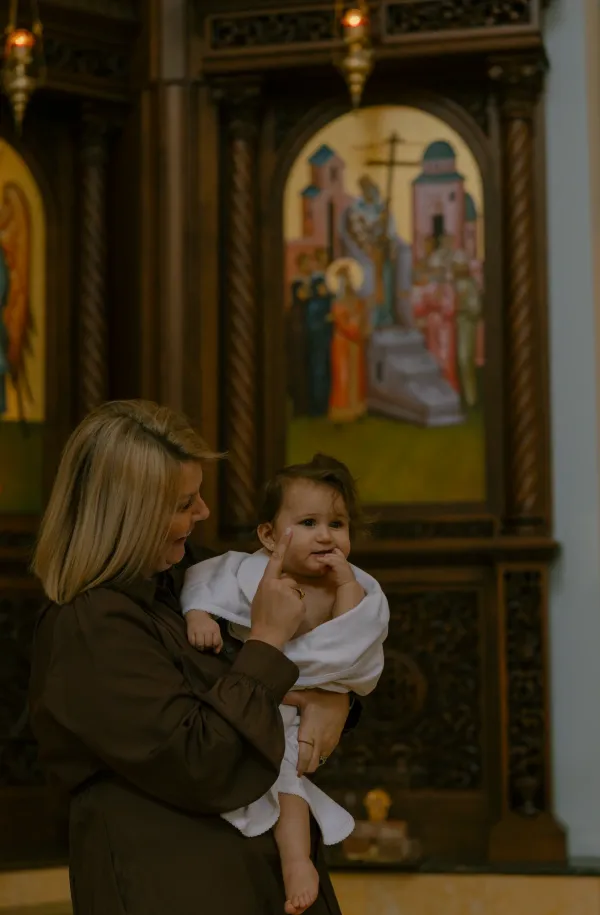Smiling woman holding a baby wrapped in white during a christening ceremony inside a church.