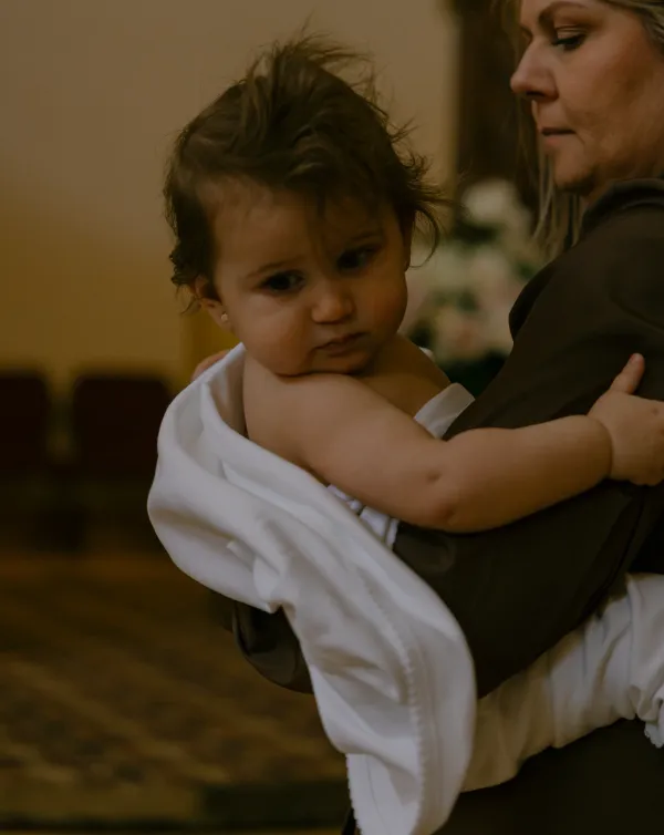 A sweet baby girl with messy hair, wrapped in white, held by a woman during a christening ceremony.
