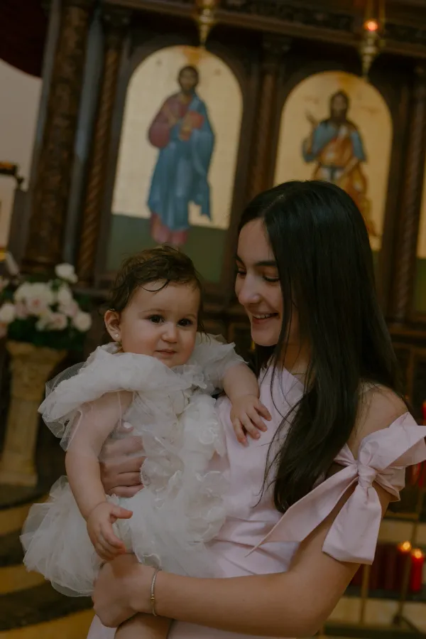 Smiling woman holding baby in white christening gown inside a church with religious icons