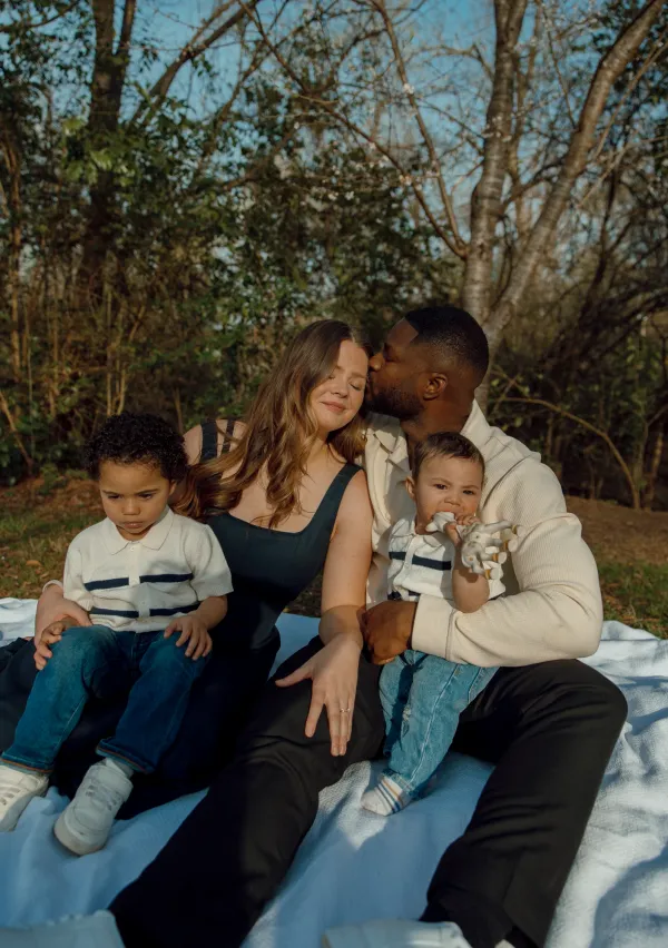 Diverse family of four on a white blanket outdoors, dad kissing mom, baby holding toy, toddler nearby.