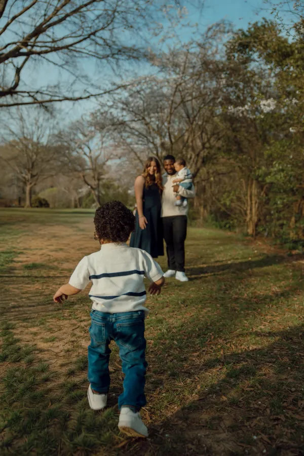 Toddler running towards parents holding baby in a Charlotte park, natural light family photo.
