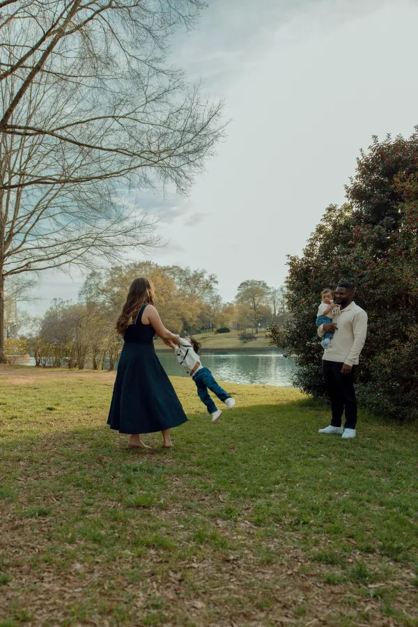 Family of four playing by a lake in Charlotte, NC. Mom swings child, dad holds baby.