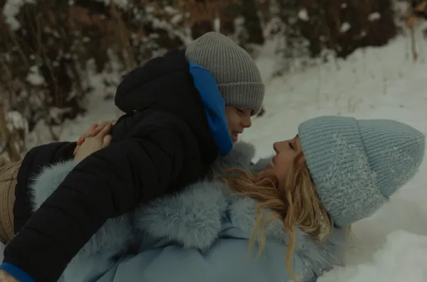 Mother and son smiling, lying in fresh snow, wearing winter hats and coats.
