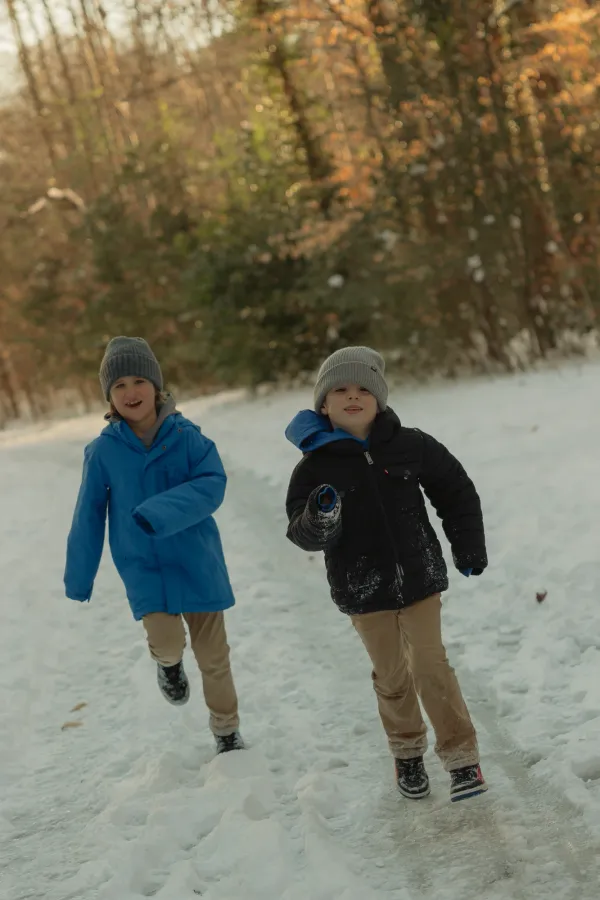 Two happy boys in winter hats and jackets running through snow, smiling at the camera.