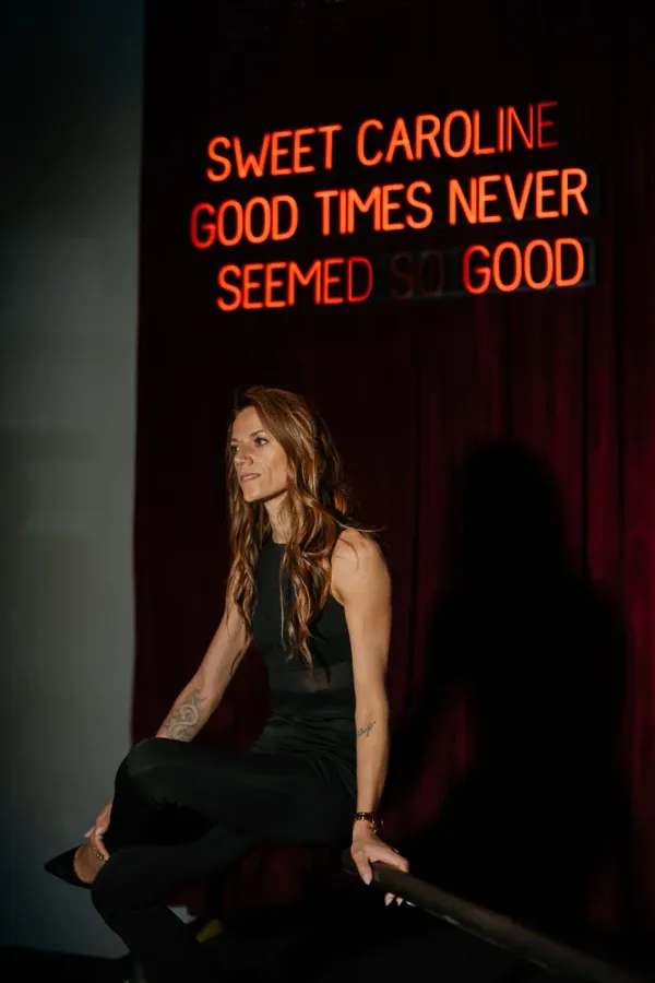 Woman in black sitting by red neon sign 'Sweet Caroline Good Times Never Seemed So Good' in Charlotte.