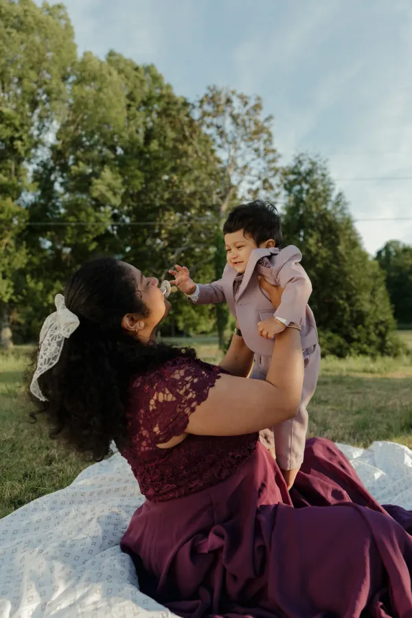 Mother in maroon dress playing with baby in lavender suit outdoors, both smiling joyfully.