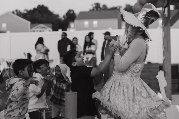Black and white photo of children laughing with face paint at an outdoor event, interacting with an adult in a large hat.