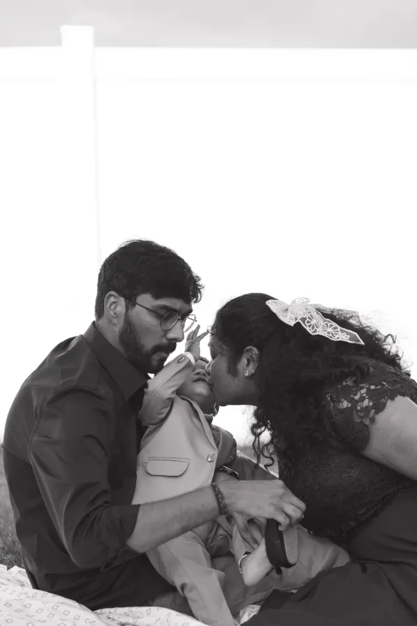 Black and white photo of parents kissing and looking at their young child outdoors.