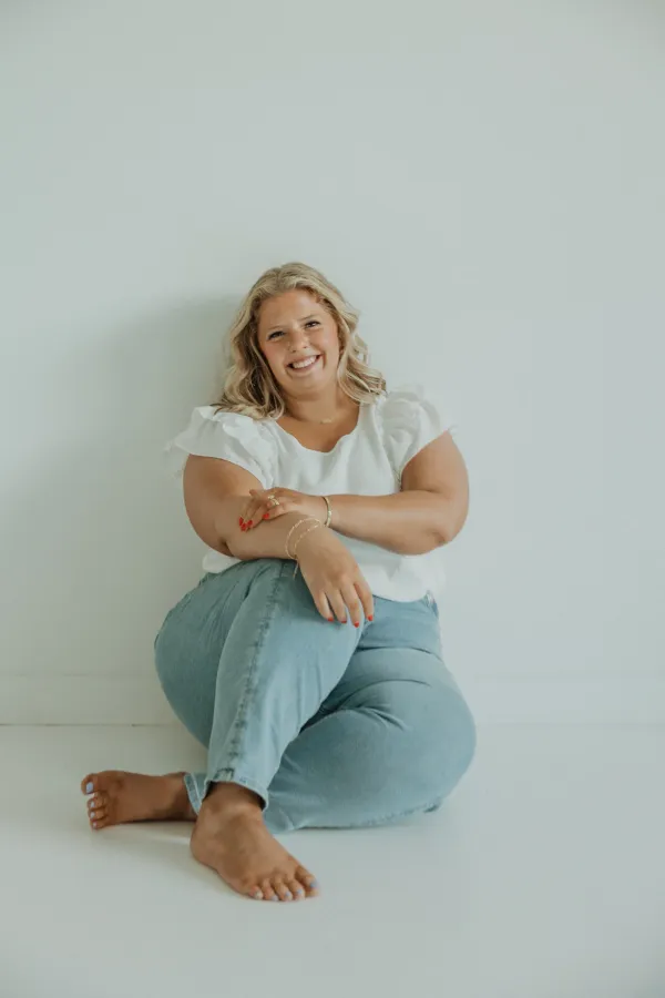 Smiling woman in white top and jeans, sitting barefoot against a white wall for a lifestyle portrait.