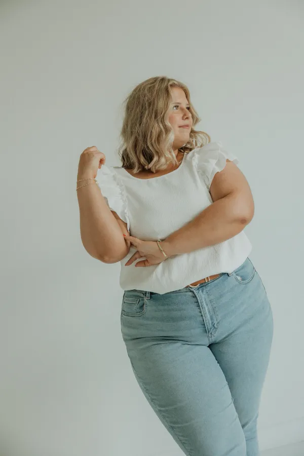 Blonde woman in white top and jeans posing confidently against a light background.