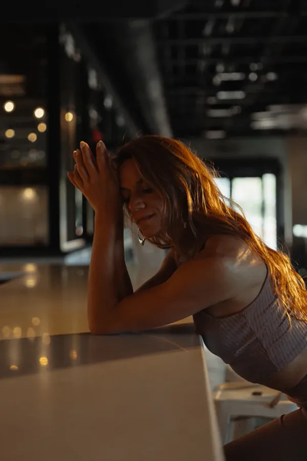 Woman with long hair, eyes closed, leaning on a bar in a cozy, warm-lit setting.