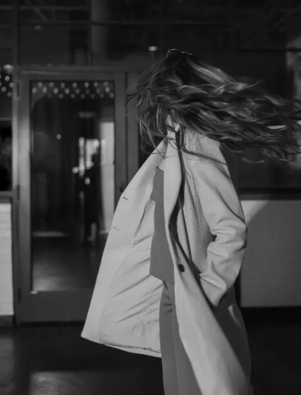 Black and white photo of a person with flowing hair and coat, walking away from a building.