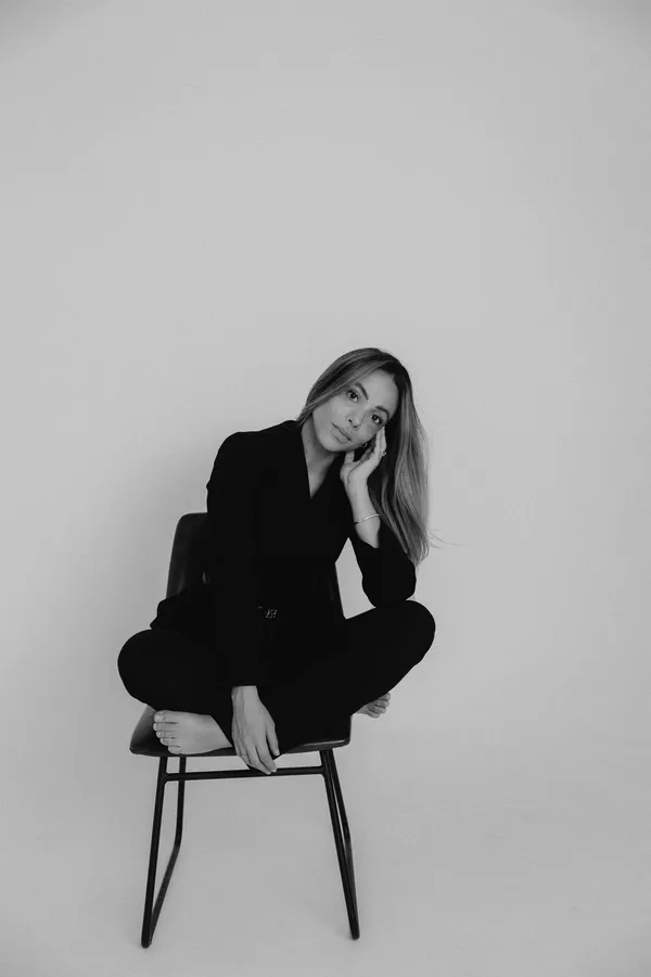 Black and white studio portrait of a professional woman with long hair, wearing a black blazer, sitting on a chair with legs crossed, looking thoughtfully at the camera. Ideal for branding and professional profiles.