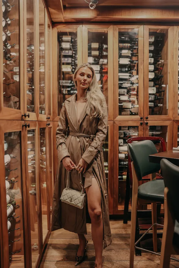 Blonde woman in a long, light brown trench coat and leopard print heels, holding a quilted white handbag, standing in a luxurious wine cellar with wooden cabinets filled with wine bottles.