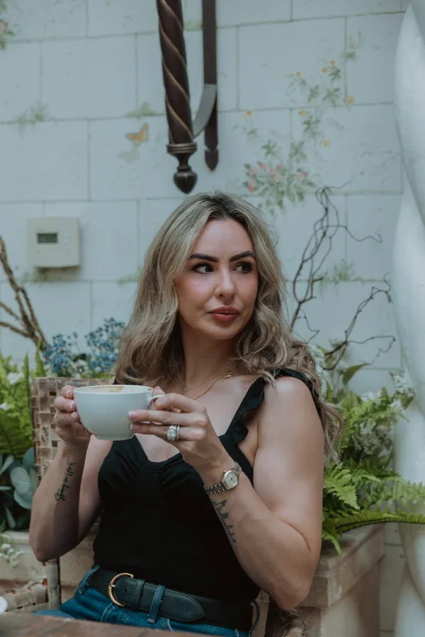 A stylish woman with blonde hair, wearing a black top and jeans, holding a white coffee cup and looking thoughtfully to the side, surrounded by lush greenery in Charlotte, NC.