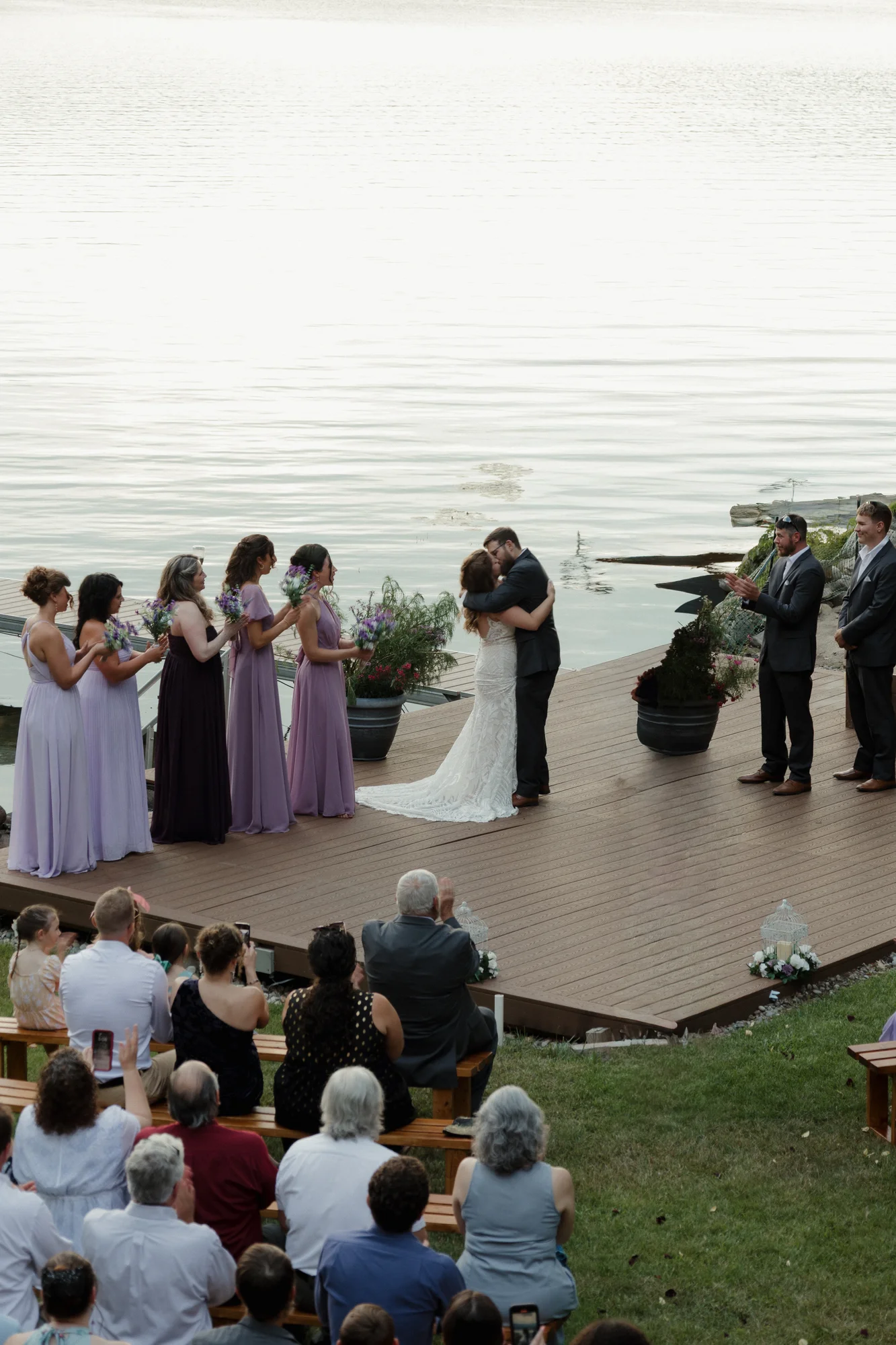 Newlyweds kissing on a wooden deck by a calm lake during their wedding ceremony, with guests watching.