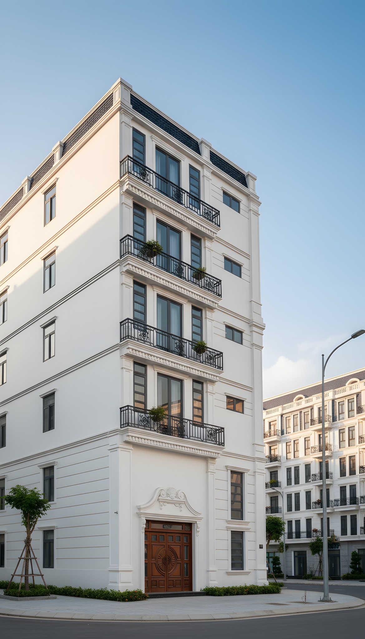 Nhà phố tân cổ điển 6 tầng nhìn từ góc nghiêng, lan can sắt uốn hoa văn, cửa gỗ tròn chạm khắc đặc sắc. Nằm trong khu đô thị mới, chụp ban ngày nắng đẹp.