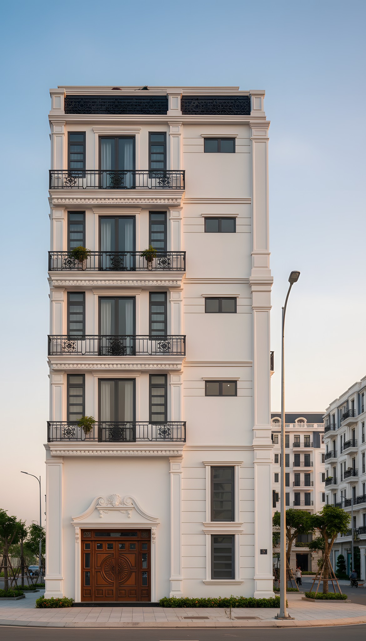 Nhà phố tân cổ điển 6 tầng nhìn từ phía trước lúc hoàng hôn, lan can sắt uốn hoa văn, cột trụ trang trí và cửa gỗ chạm khắc. Vẻ đẹp cổ điển sang trọng.