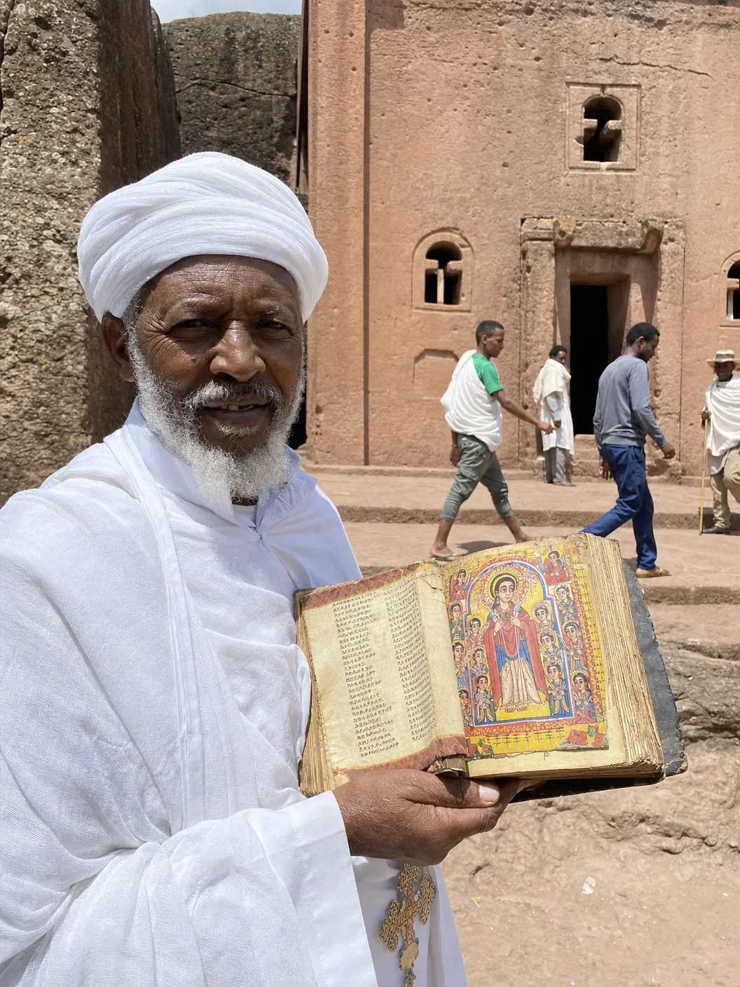 An Ethiopian Orthodox priest in white robes holding an illuminated Ge'ez Bible outside a rock-hewn church in Lalibela