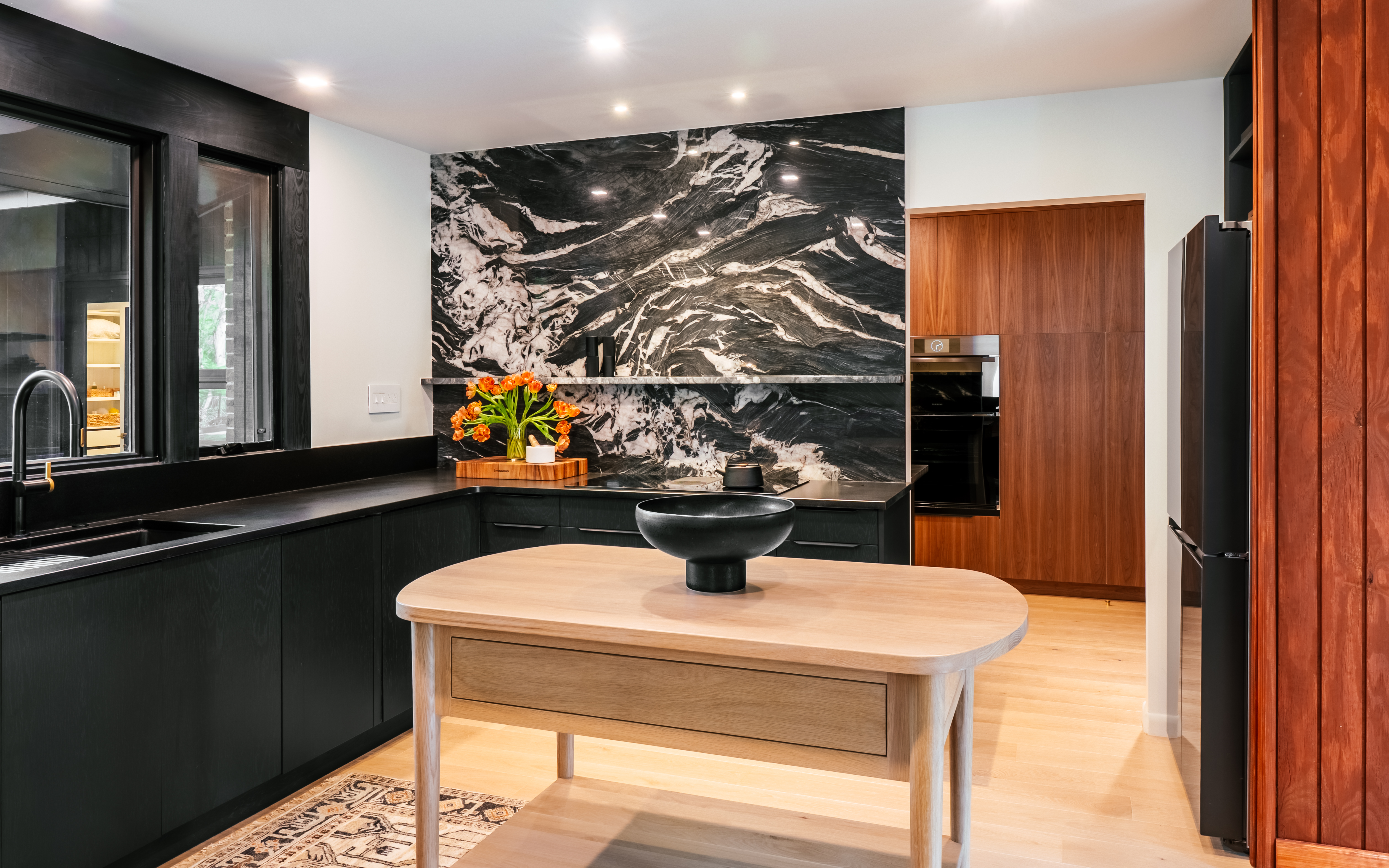 A luxurious mid-century modern kitchen features a black marble backsplash, dark cabinetry, light wood flooring, and a central light wood island with a black decorative bowl, showcasing a seamless blend of dark and light elements.