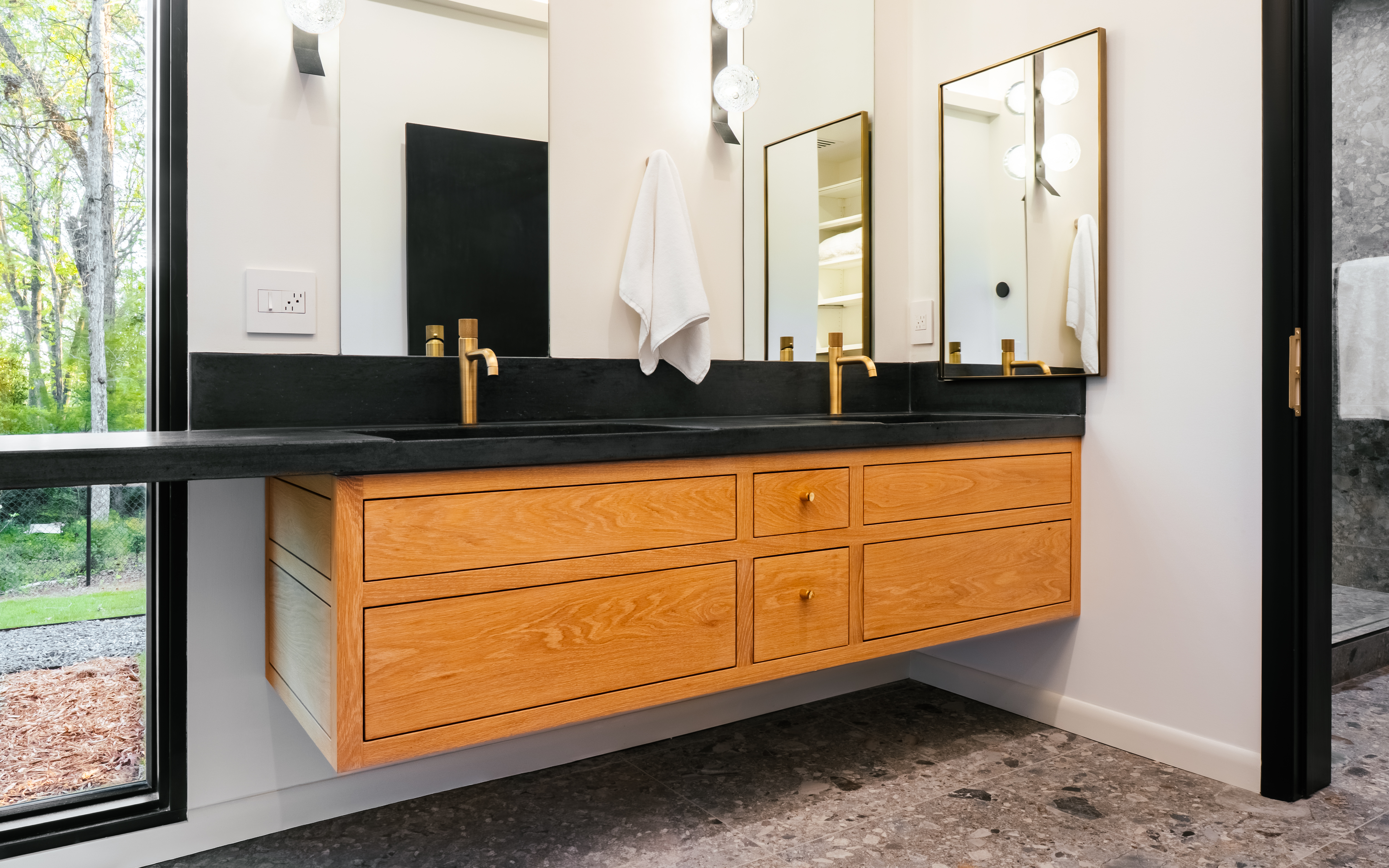 A luxurious double bathroom vanity with a floating light wood cabinet, dark stone countertop, and brass fixtures, set against a white wall with large mirrors reflecting a lush outdoor view.