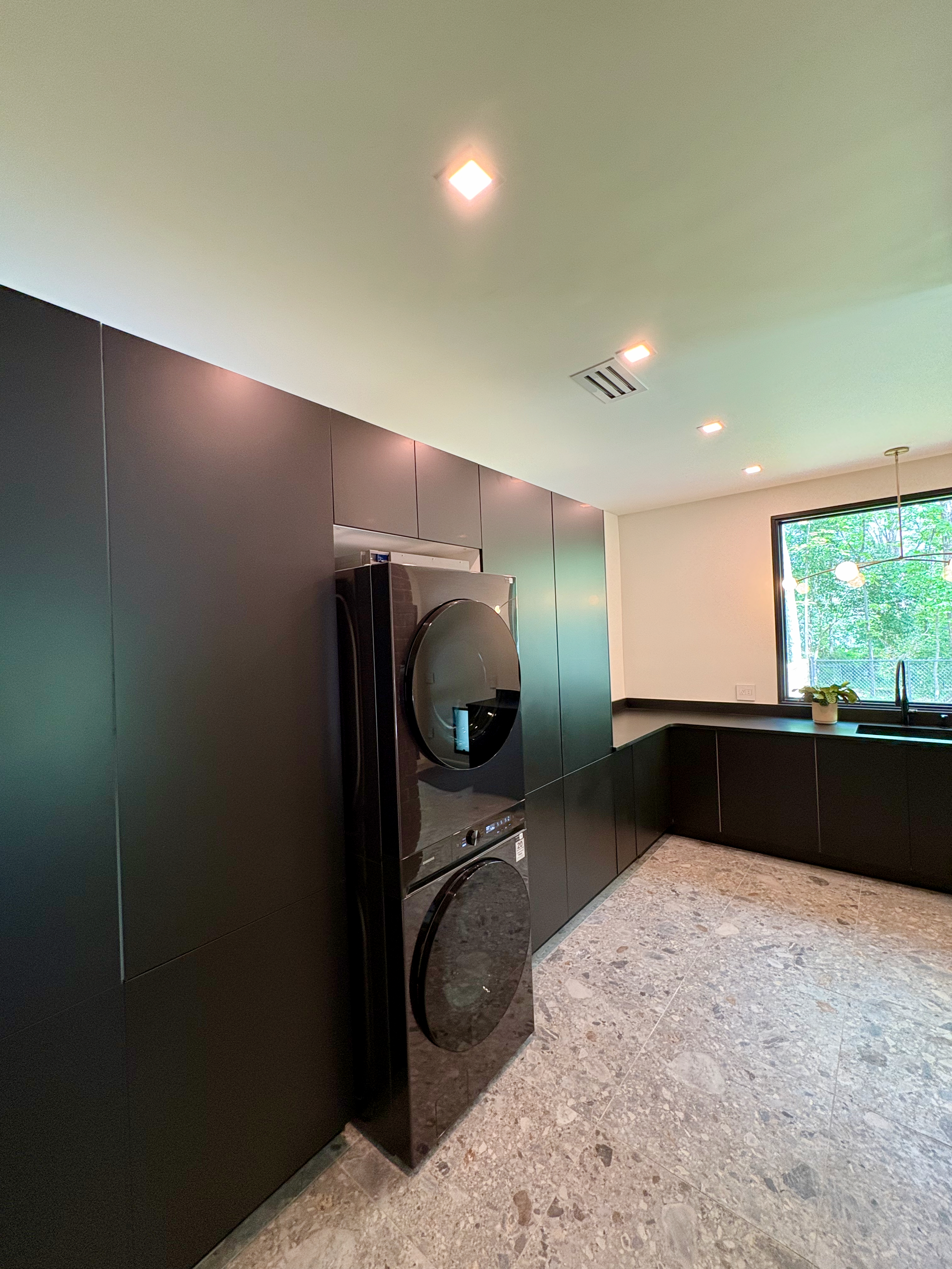 A modern laundry area featuring a stacked black washer and dryer unit, integrated flush with dark matte cabinetry, set against light speckled flooring and a large window overlooking lush greenery.