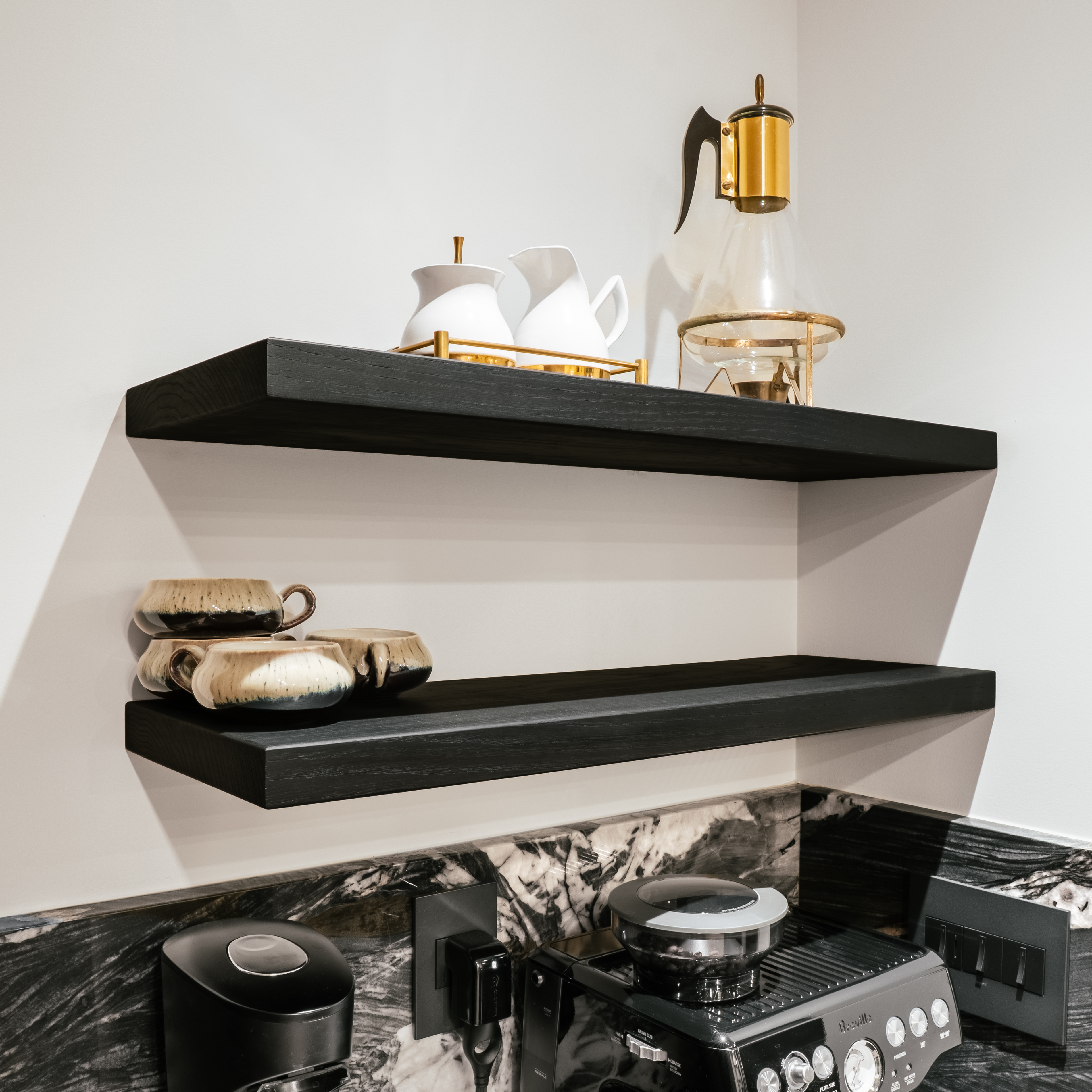 A luxurious coffee station featuring two dark wood floating shelves against a light wall, above a black marble countertop with white veining. The shelves hold stylish ceramic cups and white pitchers, while a black espresso machine and coffee grinder sit on the counter.