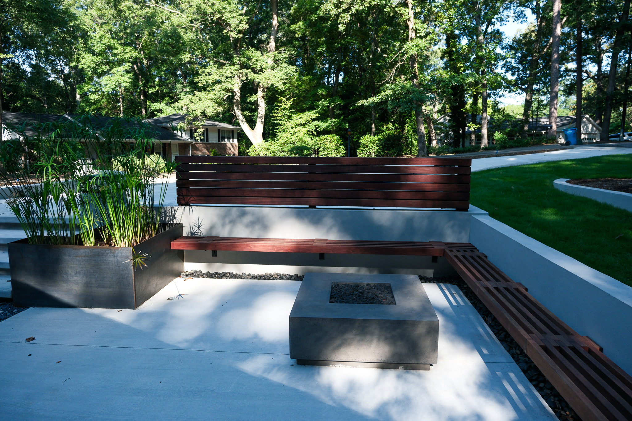 A modern outdoor patio featuring a concrete fire pit, built-in wooden benches, and a large planter with tall green plants, all set against a backdrop of dense trees and a distant mid-century modern home.
