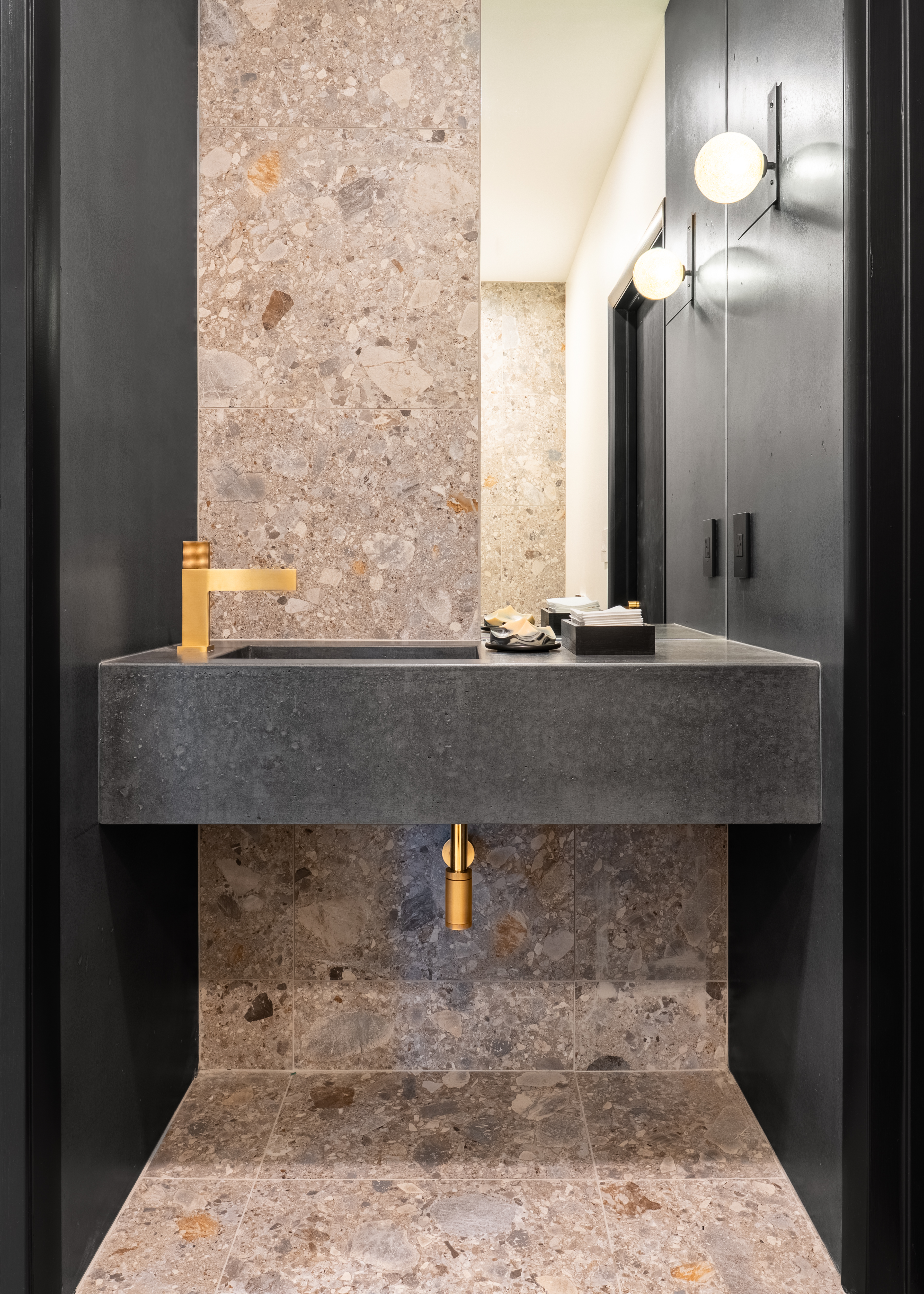 A luxurious modern powder room with a dark concrete floating vanity, a sleek gold faucet, and walls and floor clad in light brown and grey terrazzo-style tiles, illuminated by two spherical wall sconces.