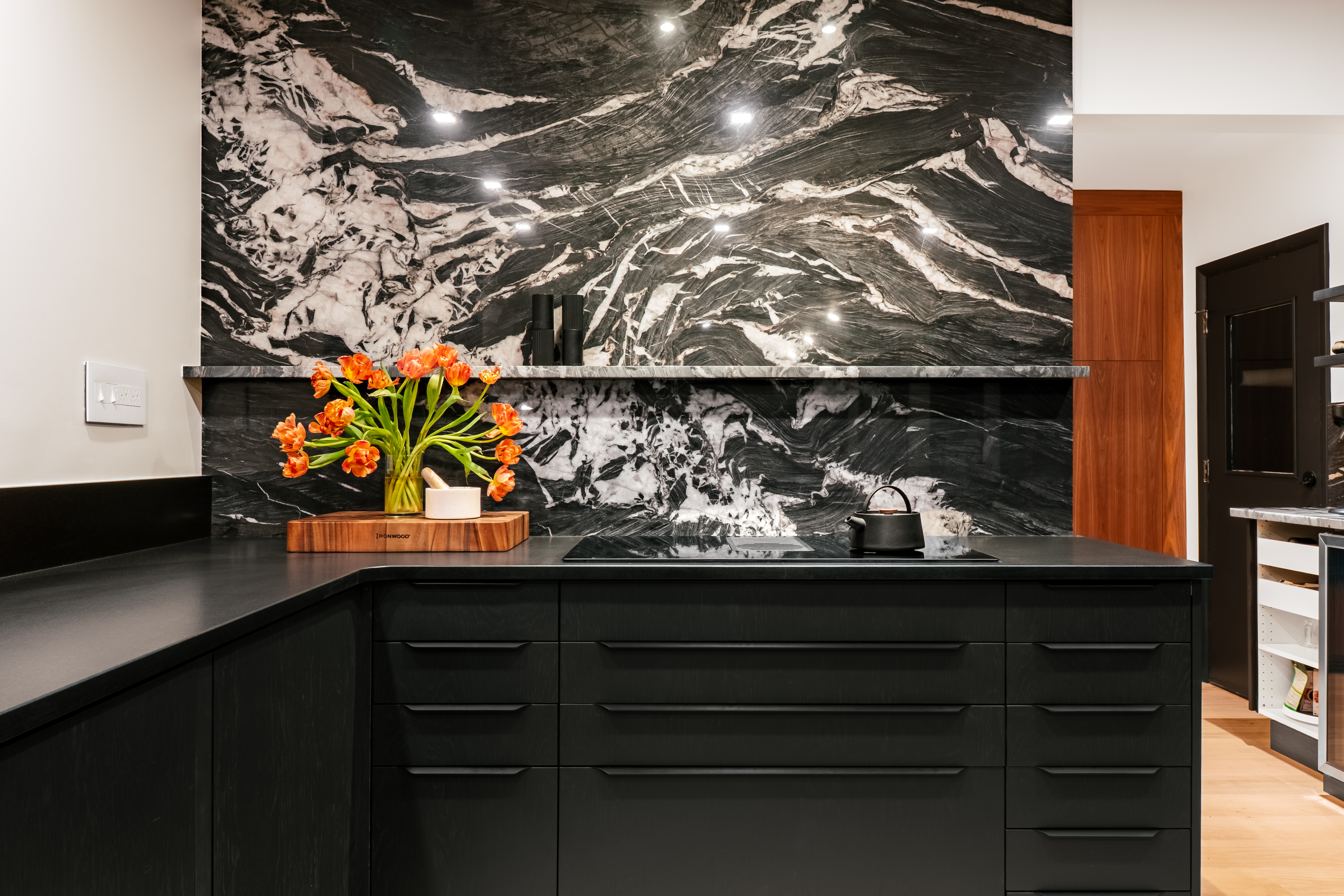 A luxurious kitchen featuring a striking black and white marble backsplash and dark, minimalist cabinetry, with a black countertop holding a vase of orange tulips and a black teapot.