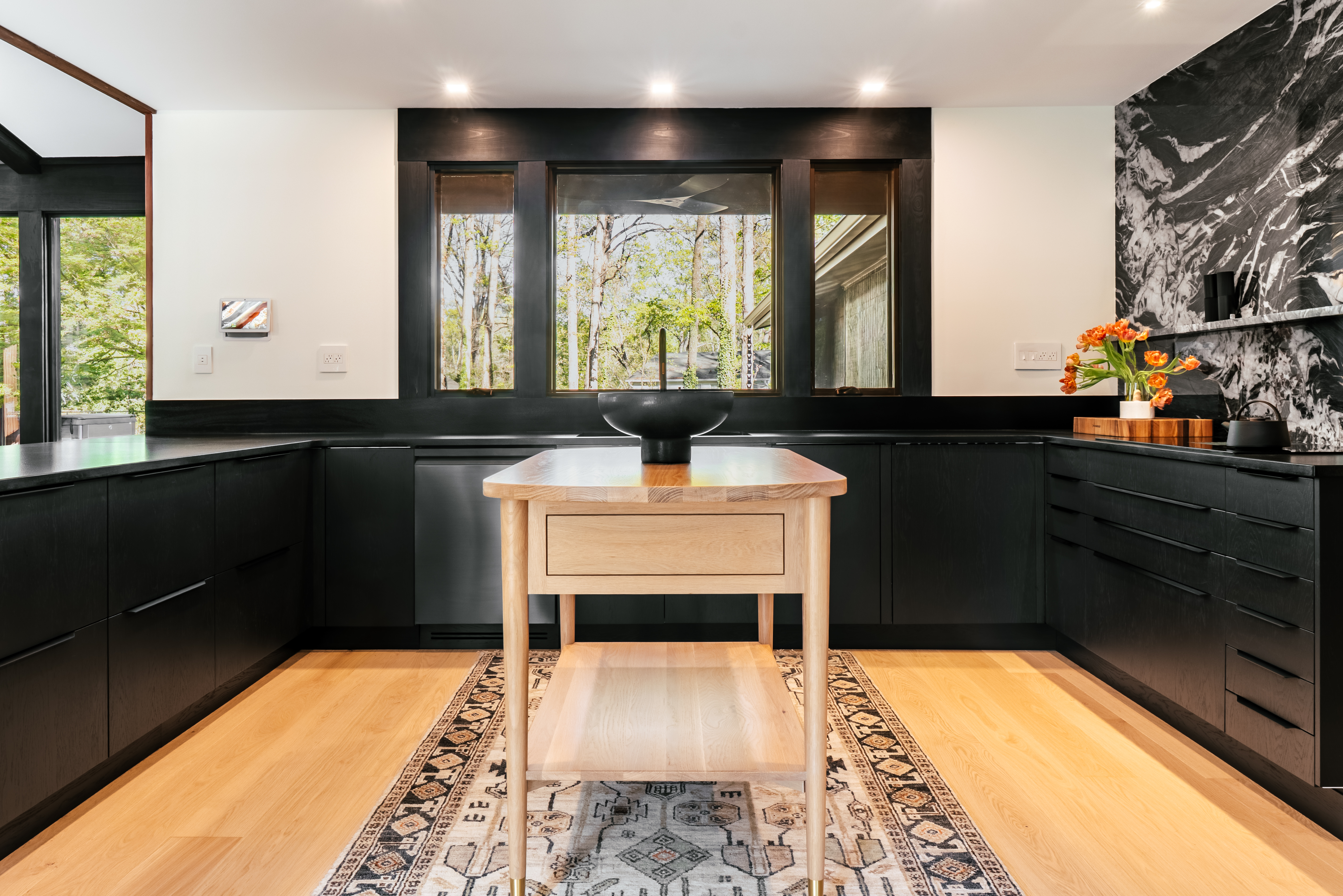 A modern kitchen featuring dark wood cabinetry, black countertops, and a striking black and white marble backsplash, complemented by a light wood island and flooring, with large windows overlooking a verdant forest.