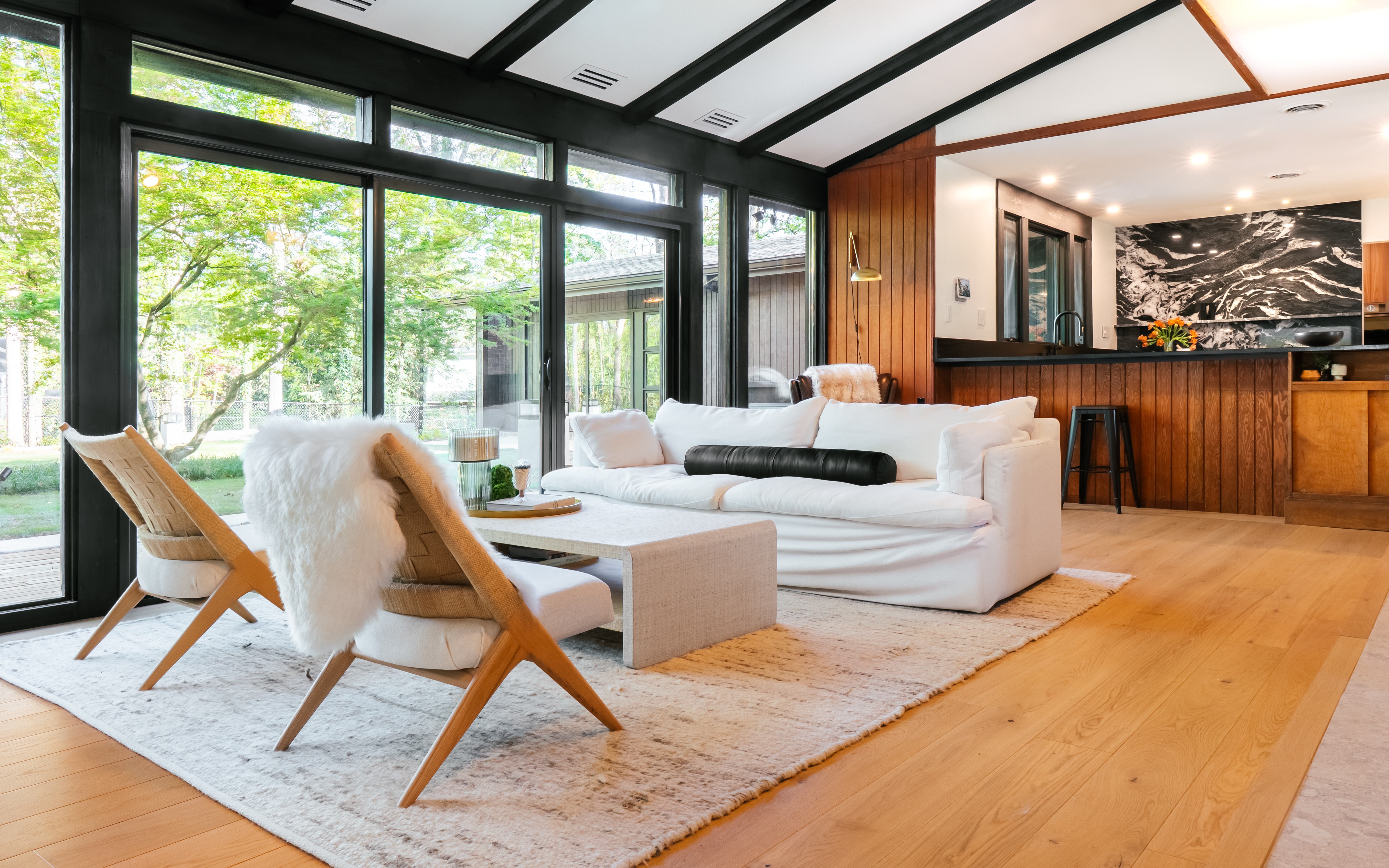 A mid-century modern living room at Ravenwood in Athens, GA, featuring a white sofa, two woven lounge chairs with white fur throws, and large black-framed windows overlooking a vibrant green outdoor landscape. The room has light wood floors, a textured rug, and a kitchen area with a black and white marble backsplash visible in the background.
