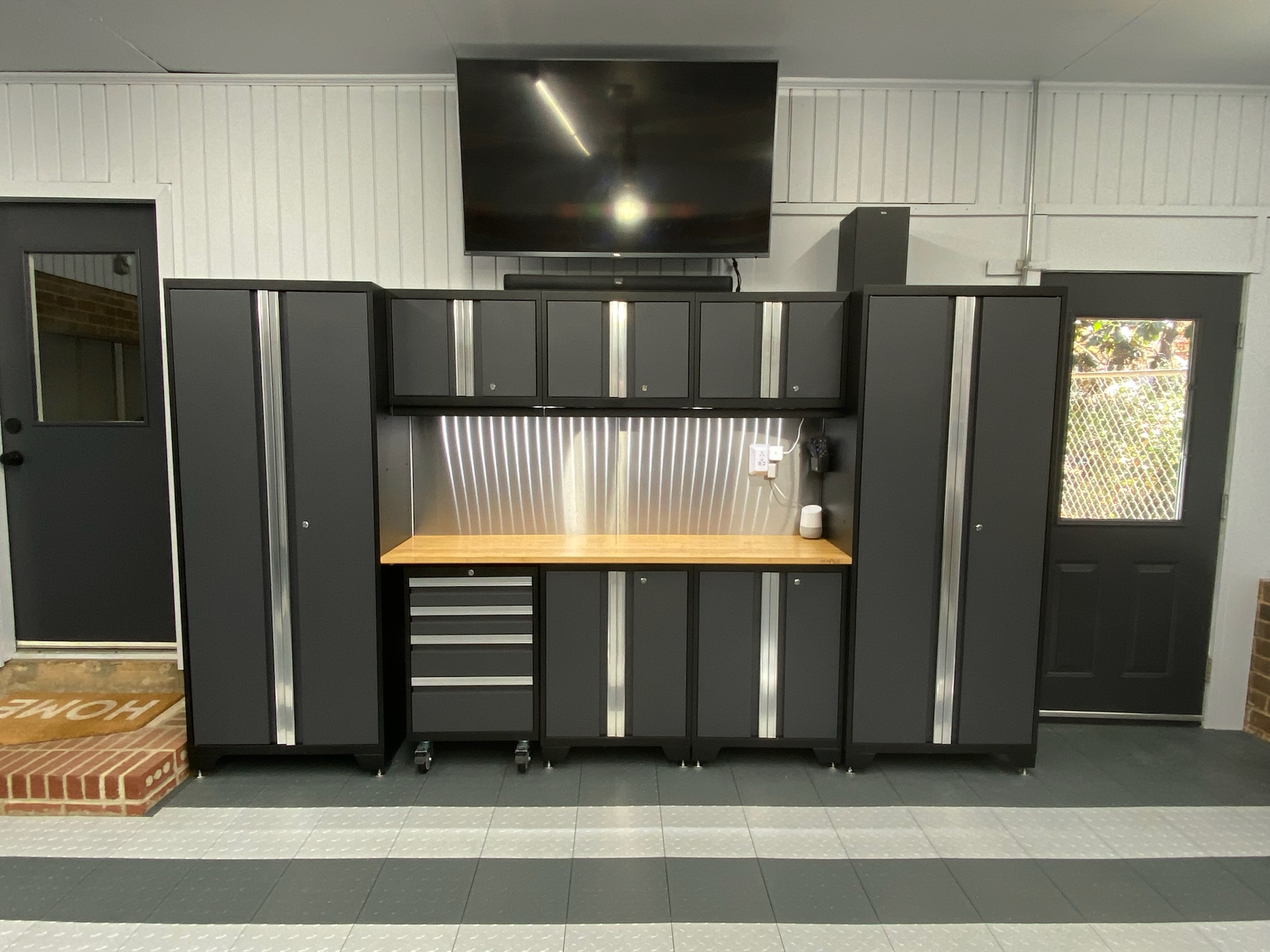 A modern garage with dark gray modular cabinets, a light wood countertop, a stainless steel backsplash, and a wall-mounted TV, all set against white paneled walls and a gray and white striped floor.