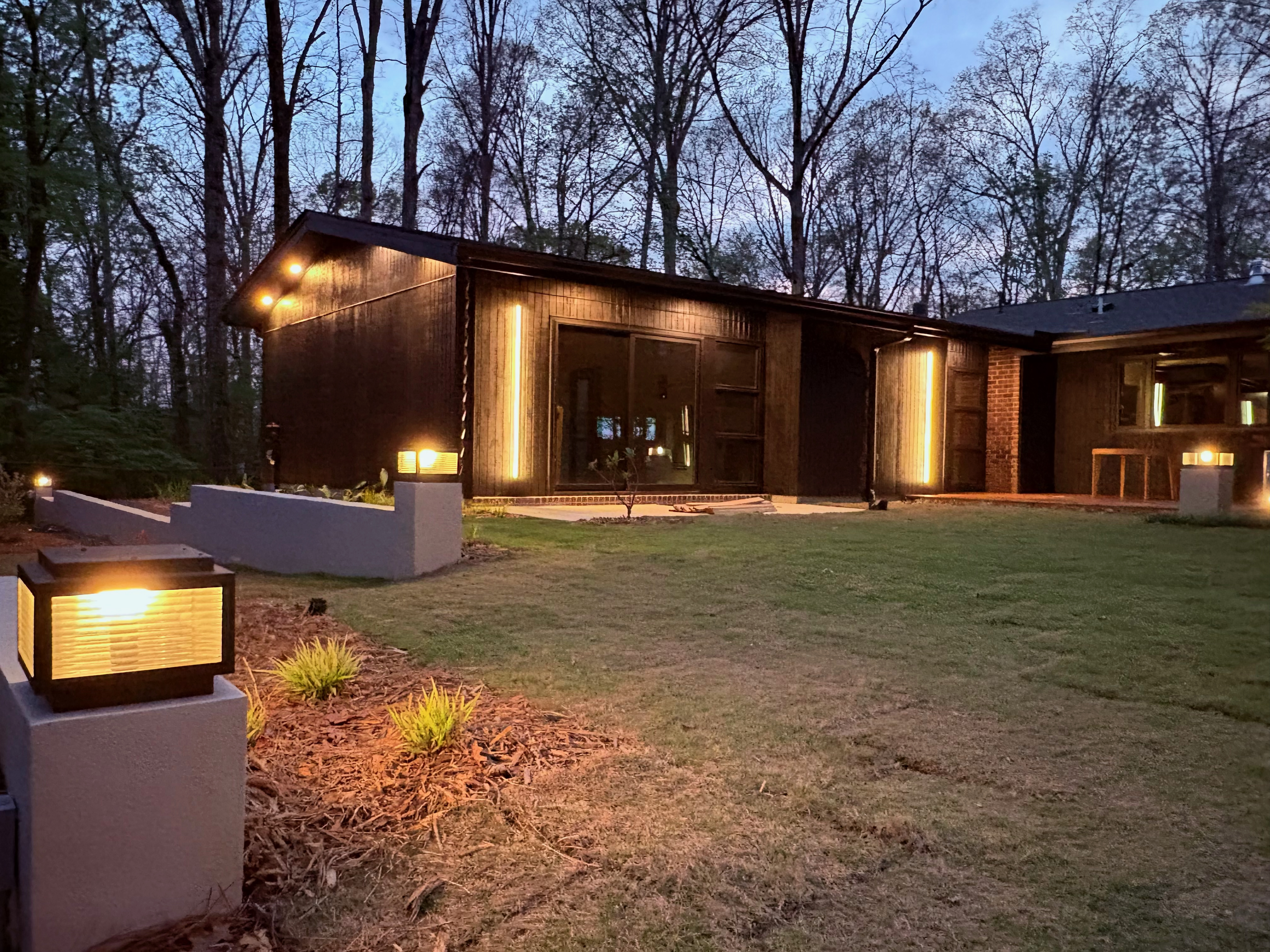 An exterior view of Ravenwood, a mid-century modern home in Athens, GA, at dusk, showcasing its dark wood facade, large windows, and illuminated vertical and path lighting against a backdrop of bare trees.