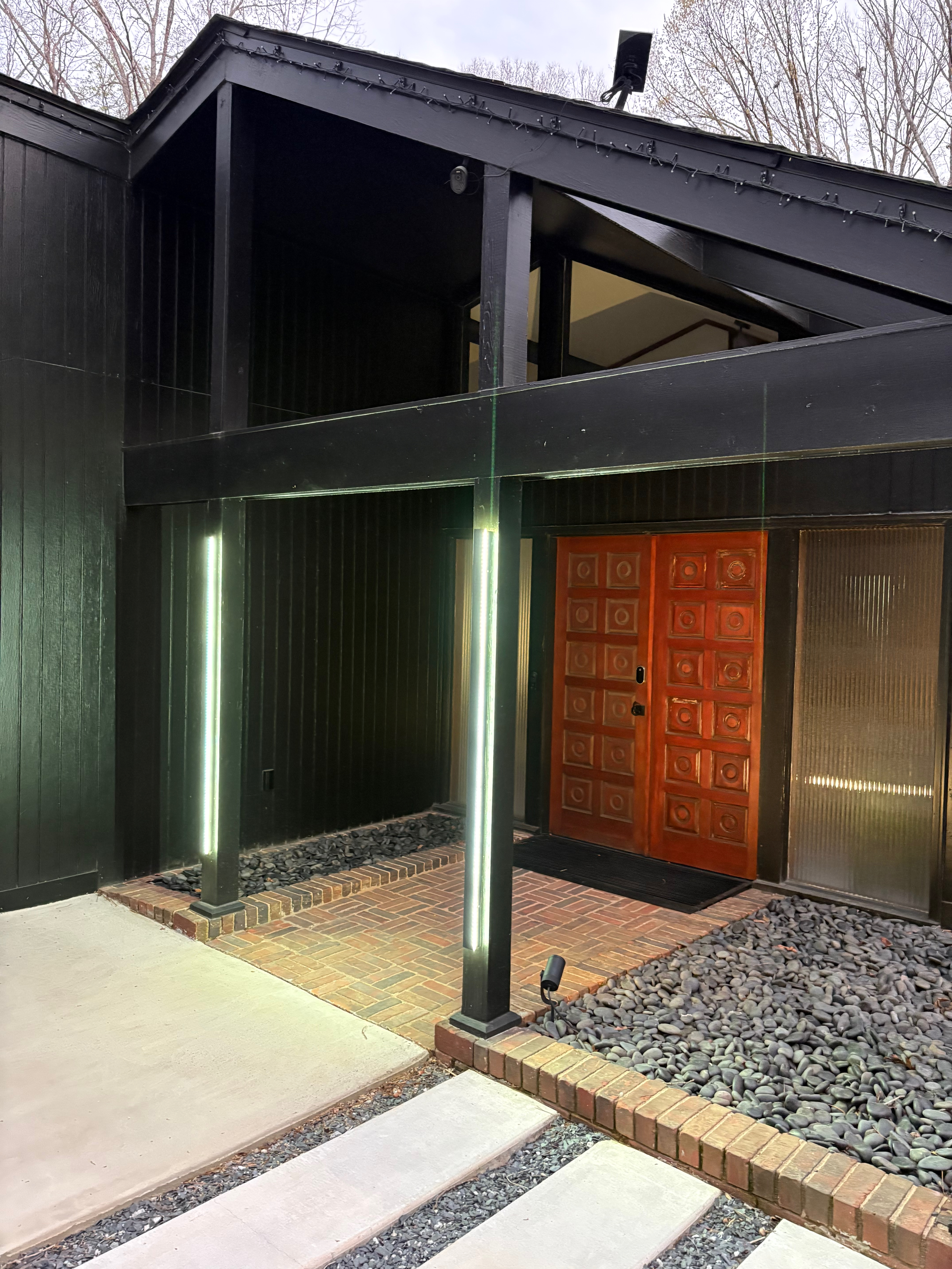 A dramatic mid-century modern home entrance features a rich orange double door with a square pattern, flanked by two vertical LED light fixtures and set against a dark, angular facade with exposed beams. The entryway is paved with brick, bordered by dark river stones, and leads to concrete stepping stones.