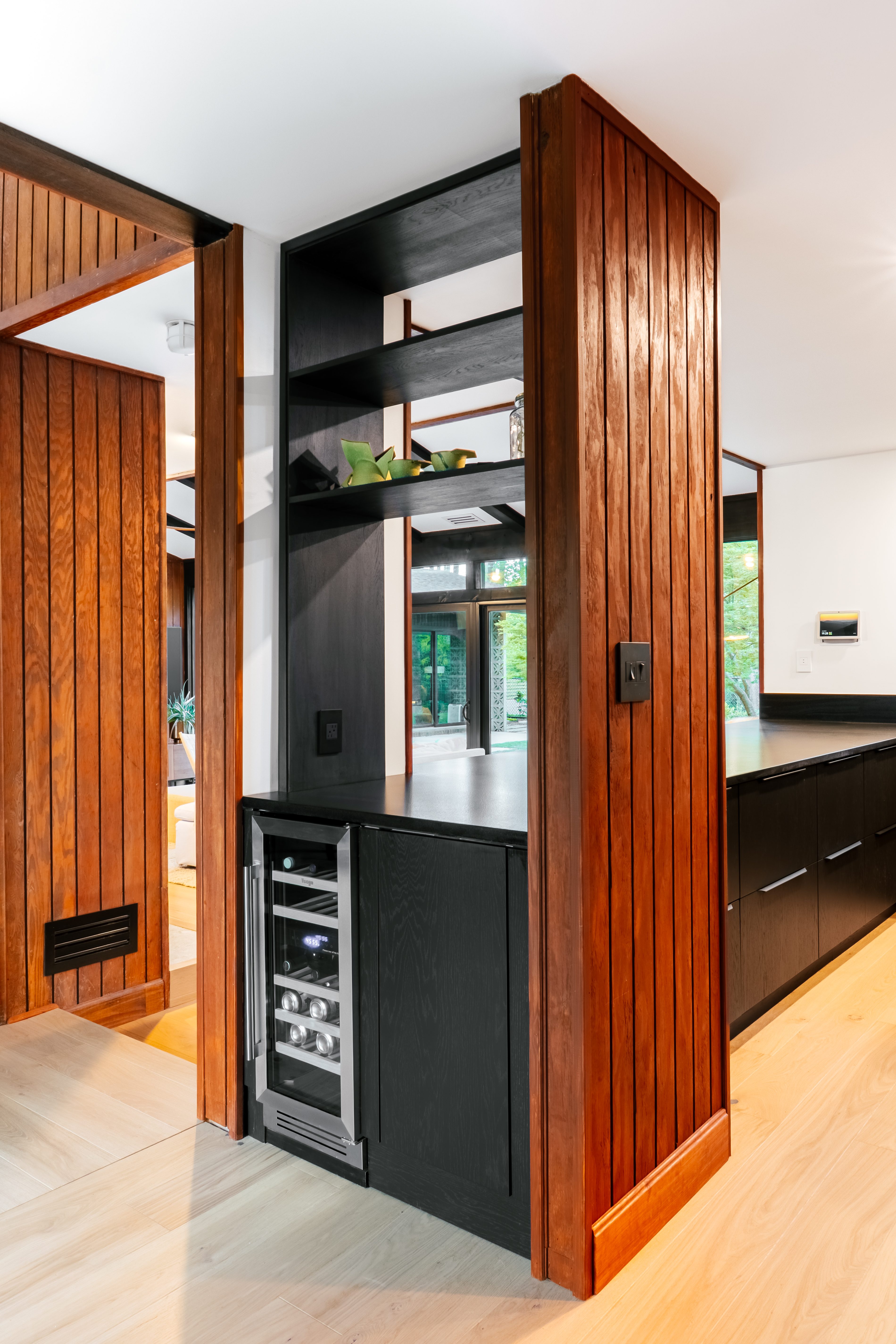 A luxurious mid-century modern interior featuring warm vertical wooden paneling contrasting with dark built-in cabinetry, an integrated wine cooler, and open shelving, all set against light wooden floors and glimpses of a lush outdoor view.