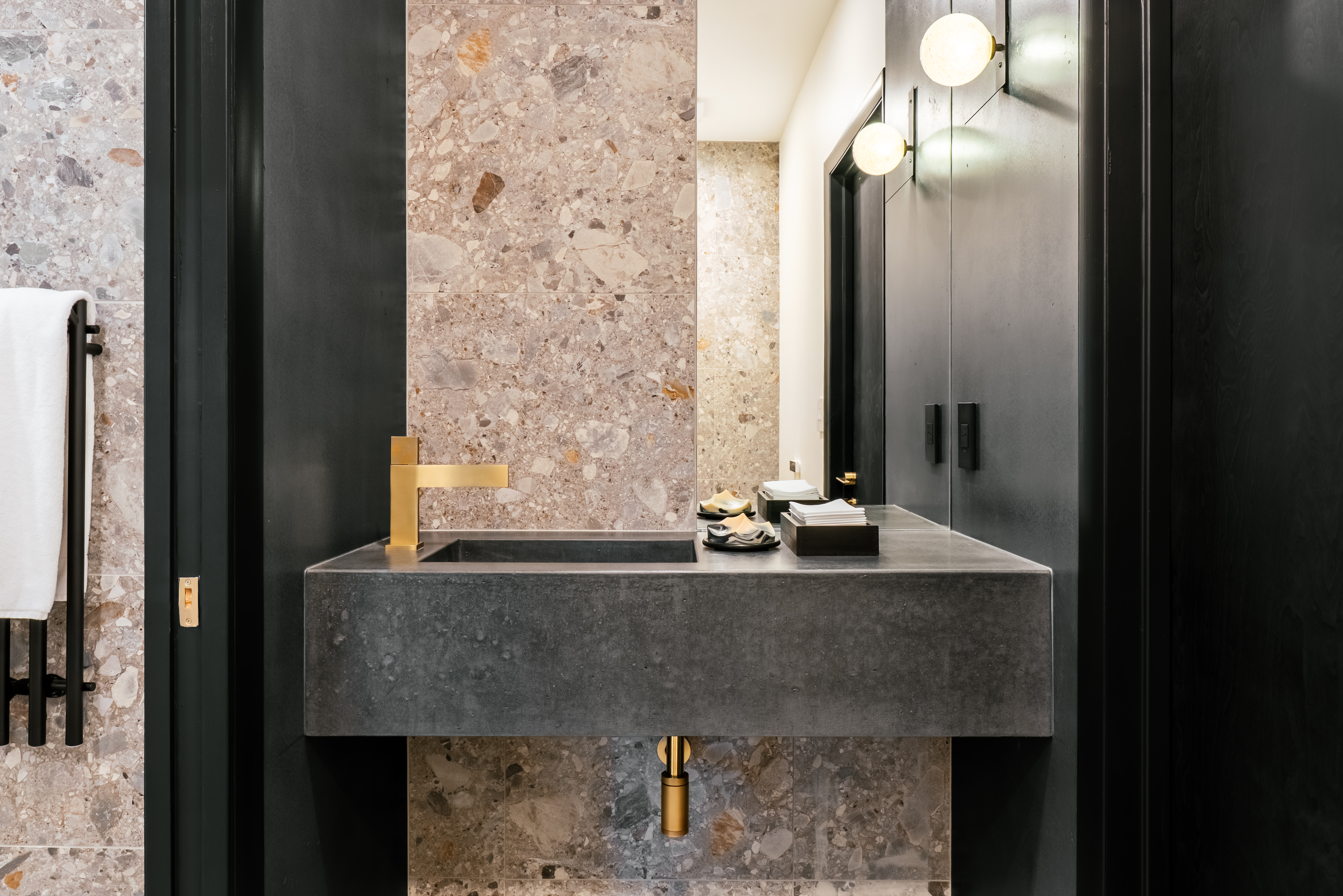 A modern bathroom features a dark gray concrete floating sink with a gold faucet, set against a wall of light brown and gray terrazzo tiles, flanked by dark wood paneling and cabinetry with integrated lighting.