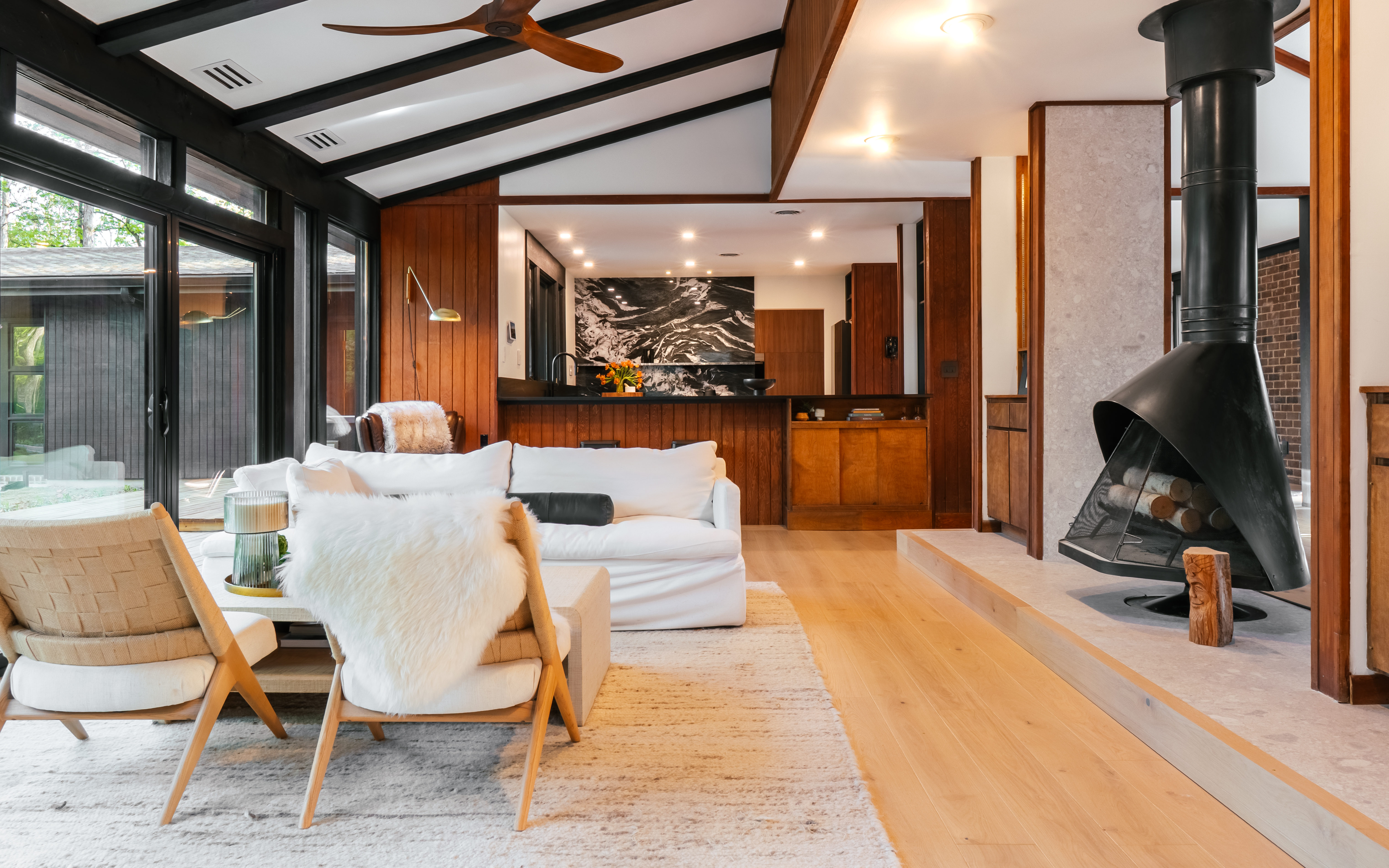 A warm, mid-century modern living room with an open layout, featuring a white sofa, two woven chairs with furry throws, a black cone-shaped fireplace, and large windows looking out to a wooded exterior. The room showcases rich wood paneling, a vaulted ceiling with dark beams, and light hardwood floors, leading into a kitchen area with a black and white marble backsplash.