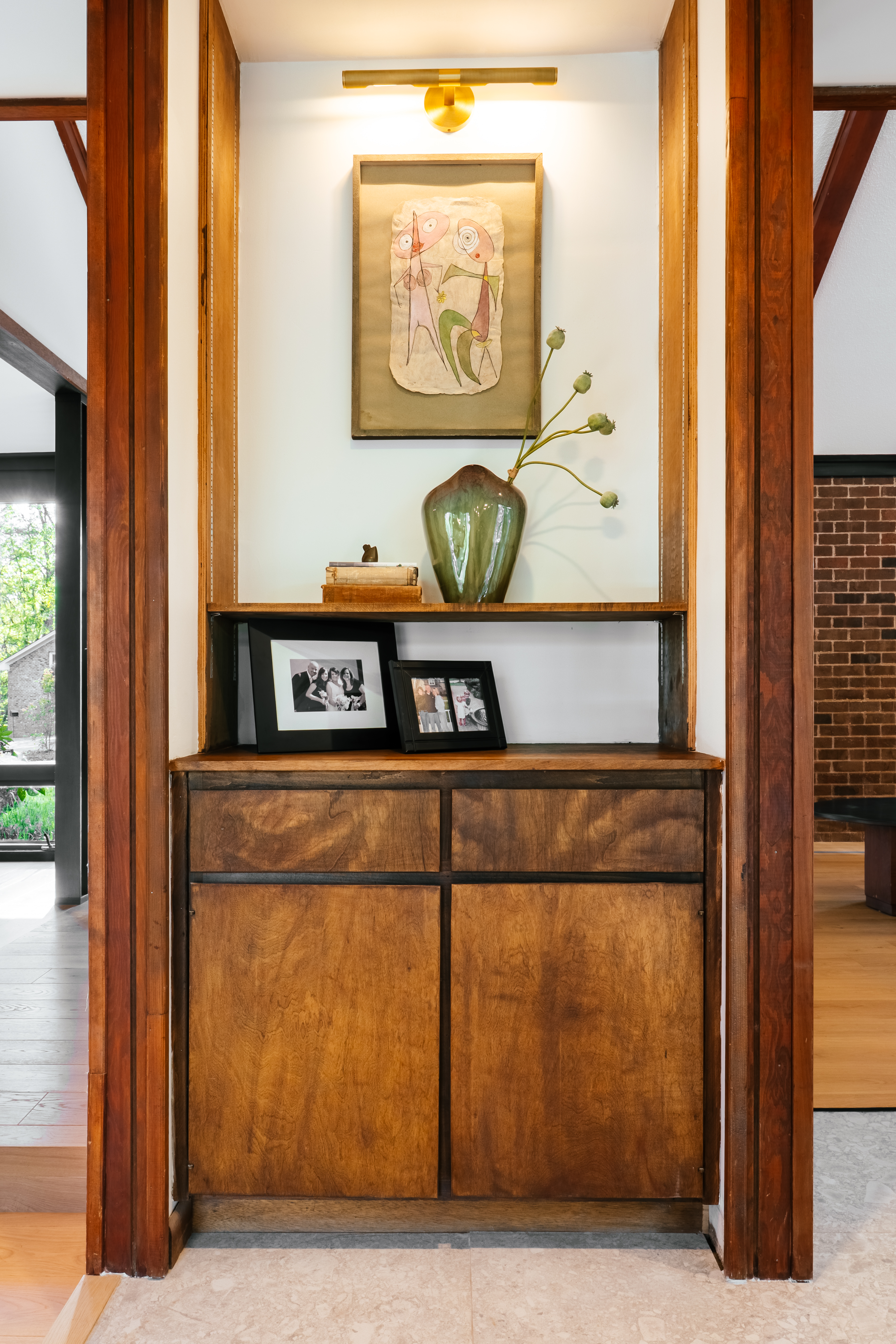 A mid-century modern built-in wooden cabinet with shelves displaying abstract artwork under a brass light, a green vase with dried poppy pods, and framed family photos, flanked by rich wooden architectural details.